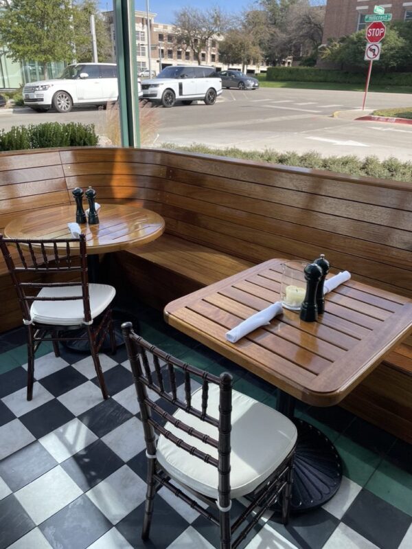 Two wooden tables with chairs and curved bench seating in a sunlit dining area next to a window with a view of the street outside.