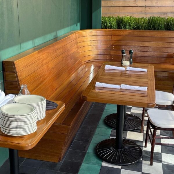 Outdoor dining area featuring a polished wooden bench and matching wooden tables set with folded napkins, along with a stack of plates on the side.