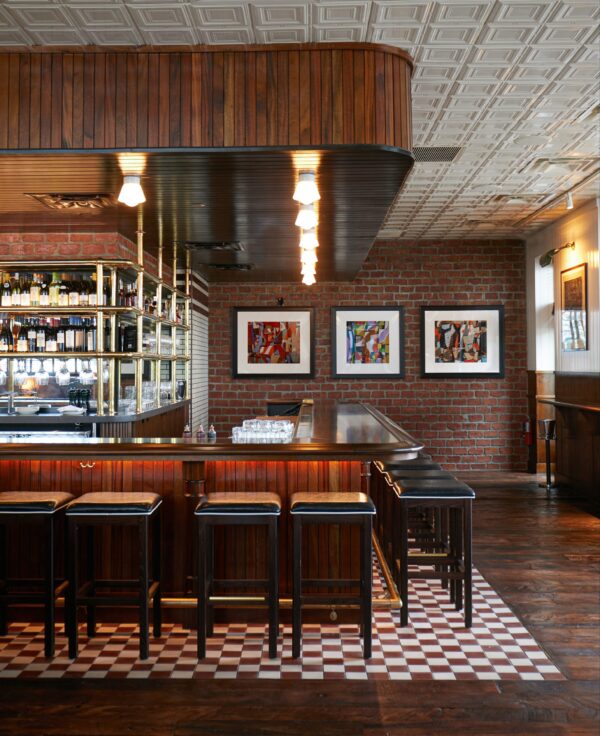 Stylish bar area with wooden paneling, checkered floor tiles, and framed abstract art on brick walls, creating a cozy yet modern atmosphere.