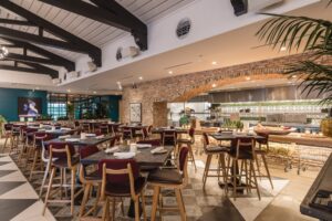 Modern restaurant interior with an open kitchen framed by a large brick arch, featuring hardwood tables with SouthLoft 260 Series commercial bases, red upholstered chairs, bar seating, decorative tile flooring, exposed ceiling beams, and warm ambient lighting.