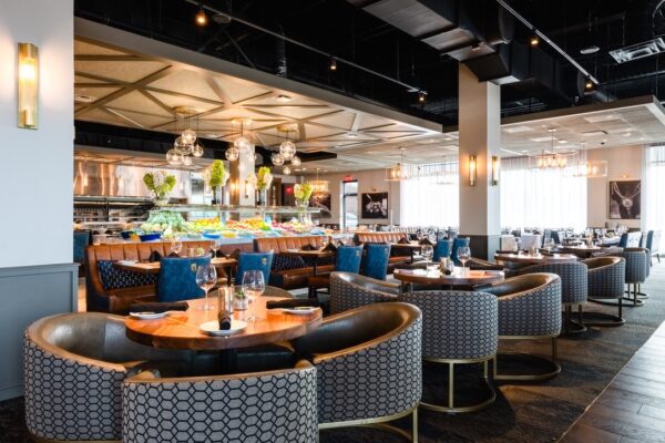 A vibrant dining area at South Loft featuring circular booth seating with patterned upholstery, blue velvet cushions, and elegant wooden tables, with a central buffet display adorned with fresh greens and fruits under modern hanging lights.