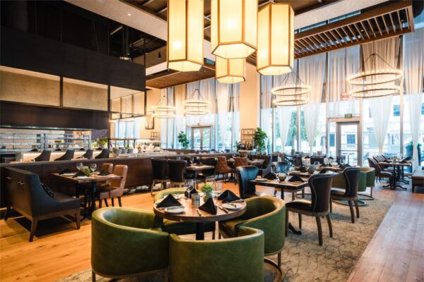 A modern dining area at South Loft featuring a mix of leather and fabric seating, warm lighting from large lantern fixtures, and a central buffet display under high ceilings with large windows.