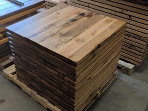 A stack of unfinished rectangular wooden tabletops on a wooden pallet, with additional wood boards and tabletops in the background.