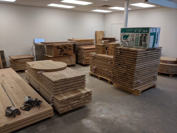 Stacks of unfinished wooden boards and tabletops in a storage room, including round and rectangular pieces, with boxes and other materials atop some stacks.