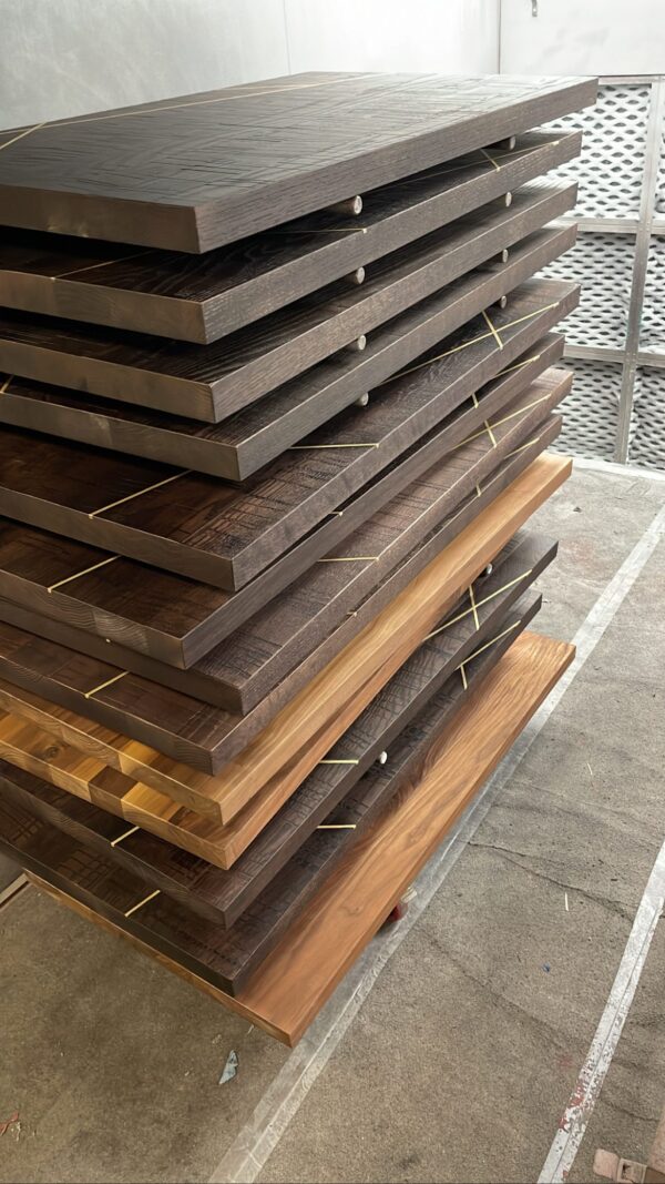 A stack of wooden tabletops of varying shades, including dark and natural wood finishes, organized in a workshop for further processing.