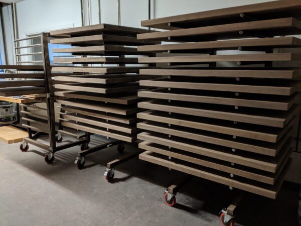 Multiple stacked wooden tabletops resting on mobile metal racks in a workshop.