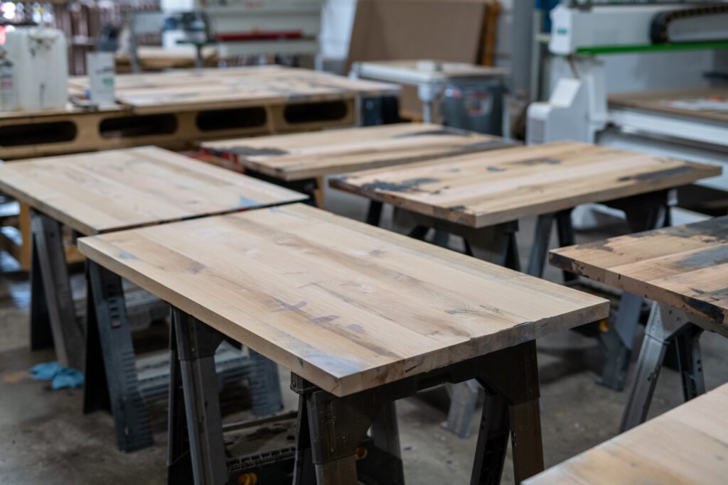 Multiple unfinished wooden tabletops are laid out on sawhorses in a workshop, undergoing preparation for further work.