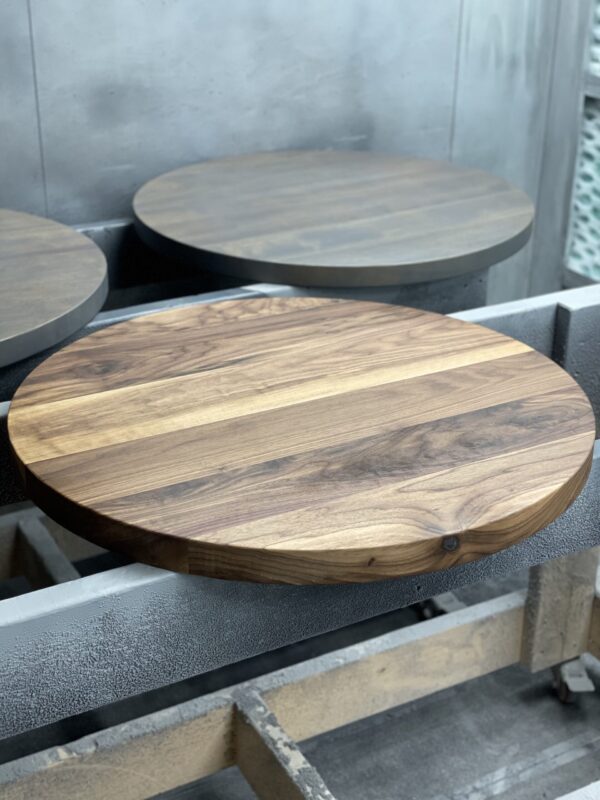 A round wooden tabletop with natural wood grain patterns is displayed in a workshop, awaiting further finishing.