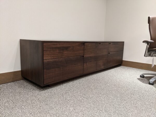 A modern dark wooden cabinet with a sleek design, set in an office space on a light gray carpet.