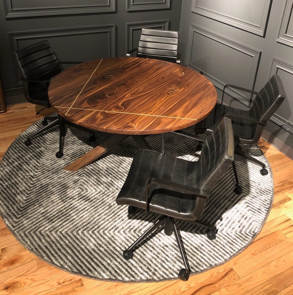 A round wooden table with intricate grain patterns and subtle brass inlays, surrounded by four black, modern office chairs on wheels, placed on a grey circular rug with a chevron pattern.