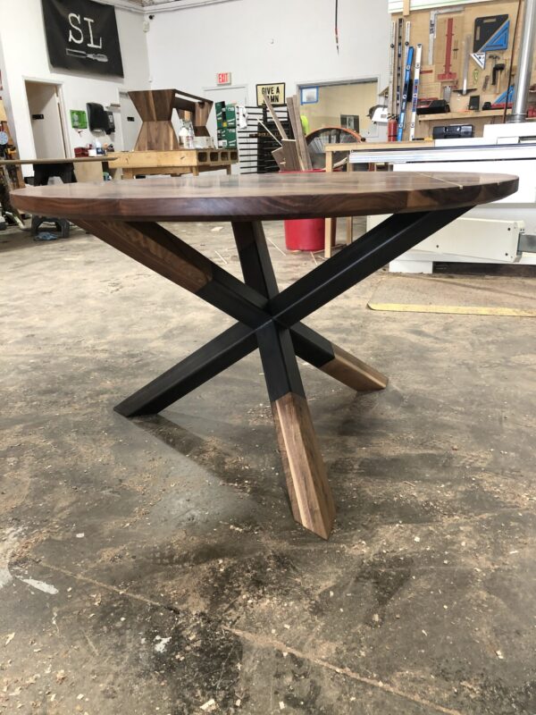 Custom round wooden table with walnut finish and modern X-shaped wood and metal base in the South Loft workshop, surrounded by woodworking tools and materials.