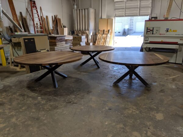 Three round wooden tables with black metal bases crafted by South Loft, displayed in a woodworking workshop with tools and materials in the background.