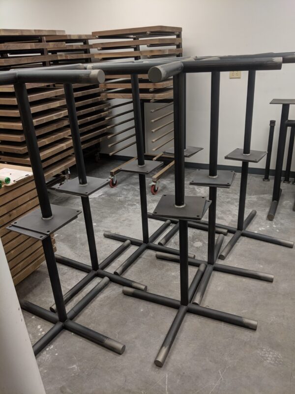 Black metal table bases with cross-shaped supports, displayed in the South Loft workshop next to stacks of wooden table tops.