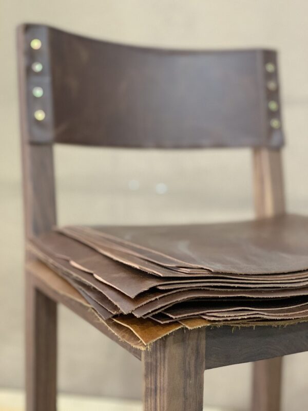 Close-up of a custom wooden chair with a leather backrest, created by South Loft, with stacked leather sheets placed on the seat.