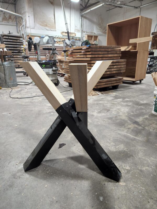 Wooden and black metal X-shaped table legs partially assembled in the South Loft workshop, with stacks of wood slabs and furniture in the background.