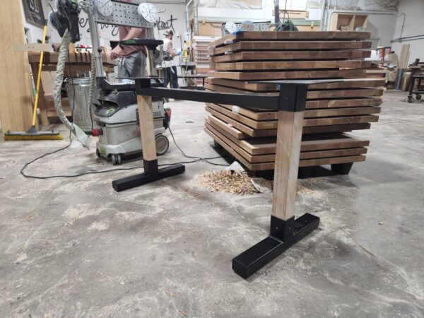 Unfinished wooden table legs with black metal supports under construction in the South Loft workshop, with wooden slabs and machinery in the background.