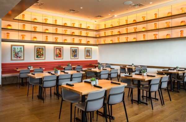 Contemporary dining room with light wooden tables, grey chairs, and red cushioned booth seating, with walls decorated by vibrant artwork and a display of orange ceramic vases.