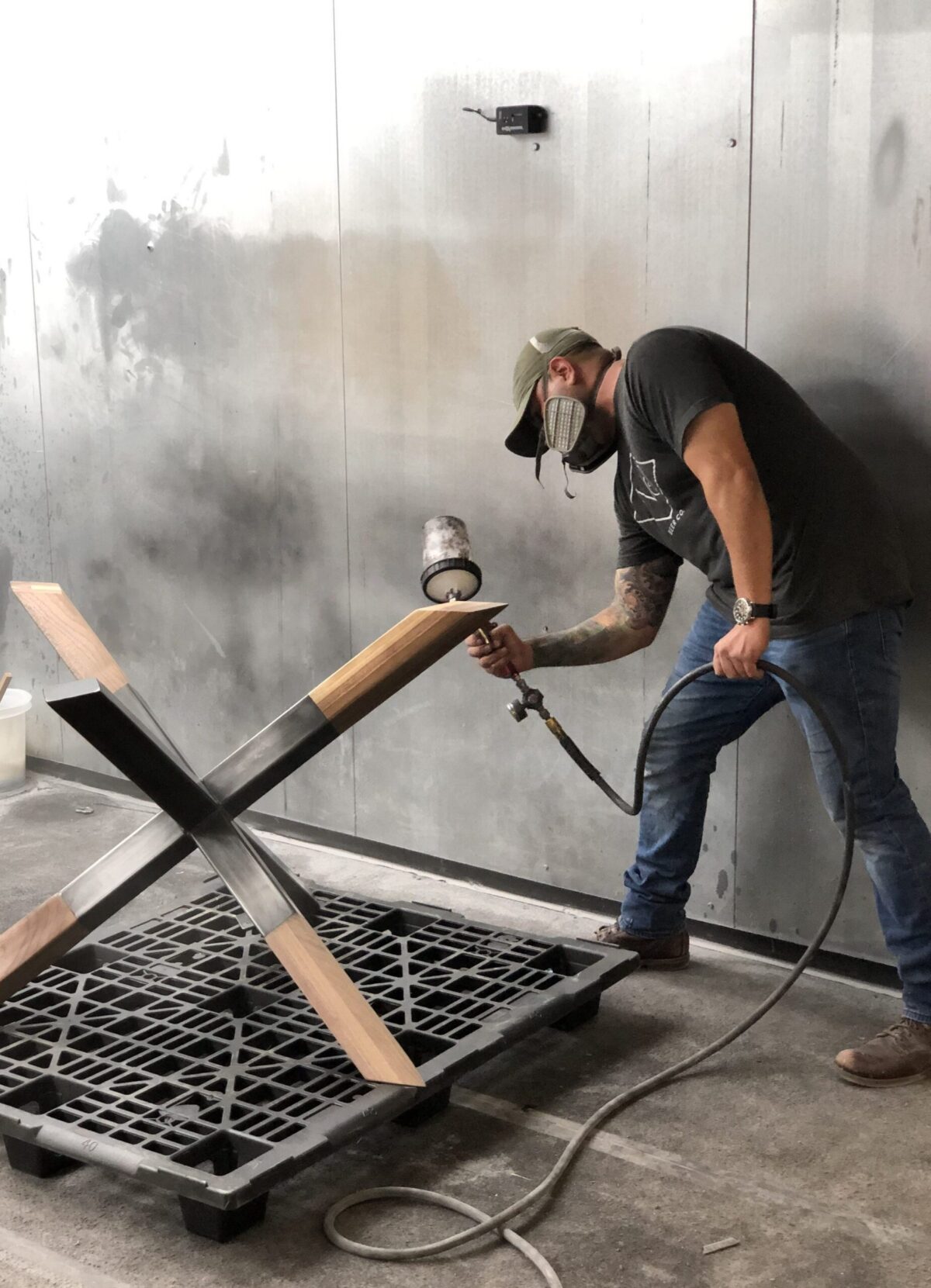 Craftsman applying finish to a wooden X-shaped table base using a spray gun in a workshop, wearing a mask and standing on a concrete floor.