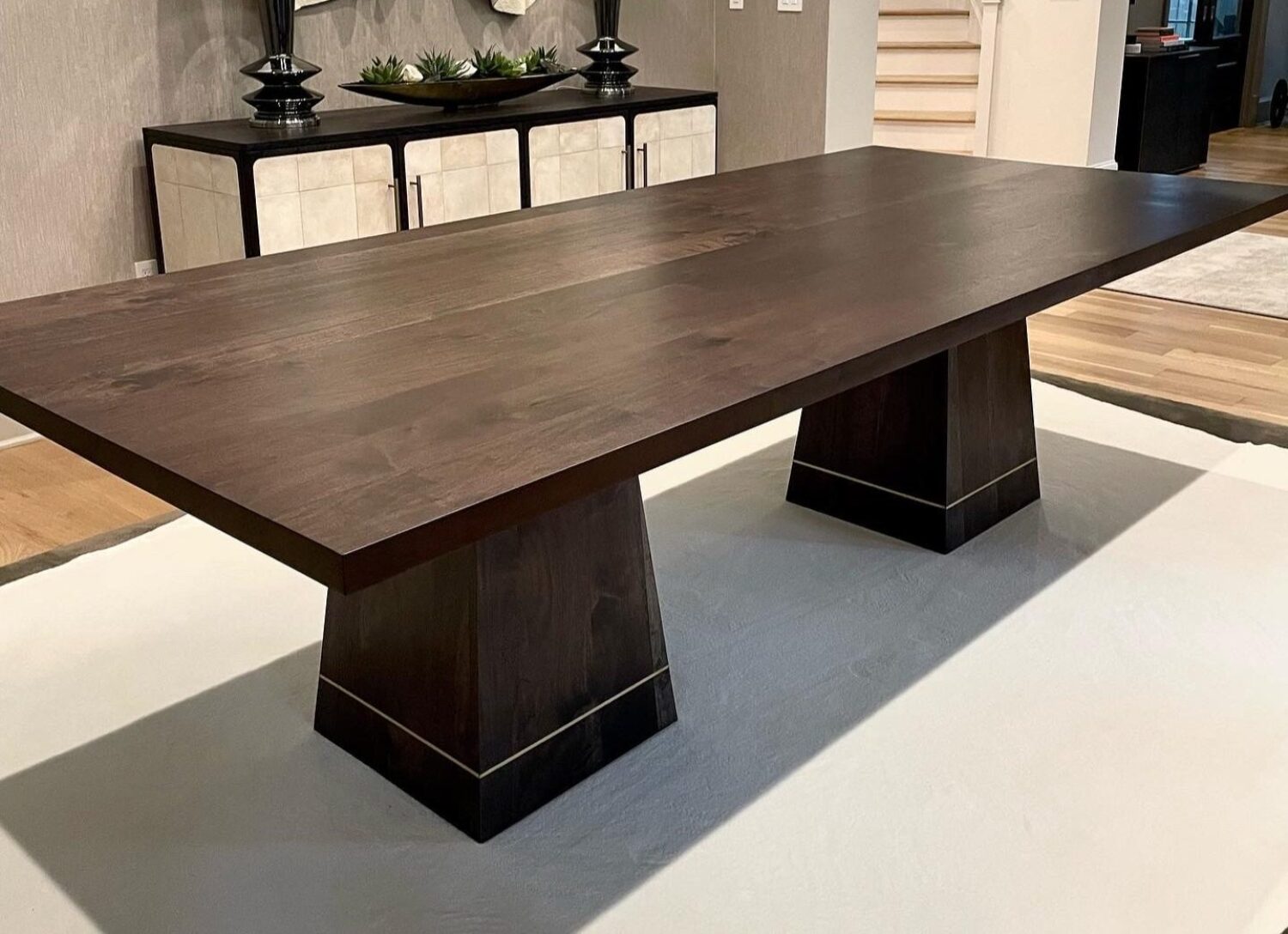 A modern dark wooden dining table with a rectangular top and two wide, tapered pedestal legs. The base of each pedestal features a subtle metallic accent line near the bottom. The table is set on a light-colored rug, with a cabinet visible in the background, adding a sleek and contemporary feel to the room.