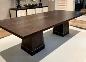 A modern dark wooden dining table with a rectangular top and two wide, tapered pedestal legs. The base of each pedestal features a subtle metallic accent line near the bottom. The table is set on a light-colored rug, with a cabinet visible in the background, adding a sleek and contemporary feel to the room.