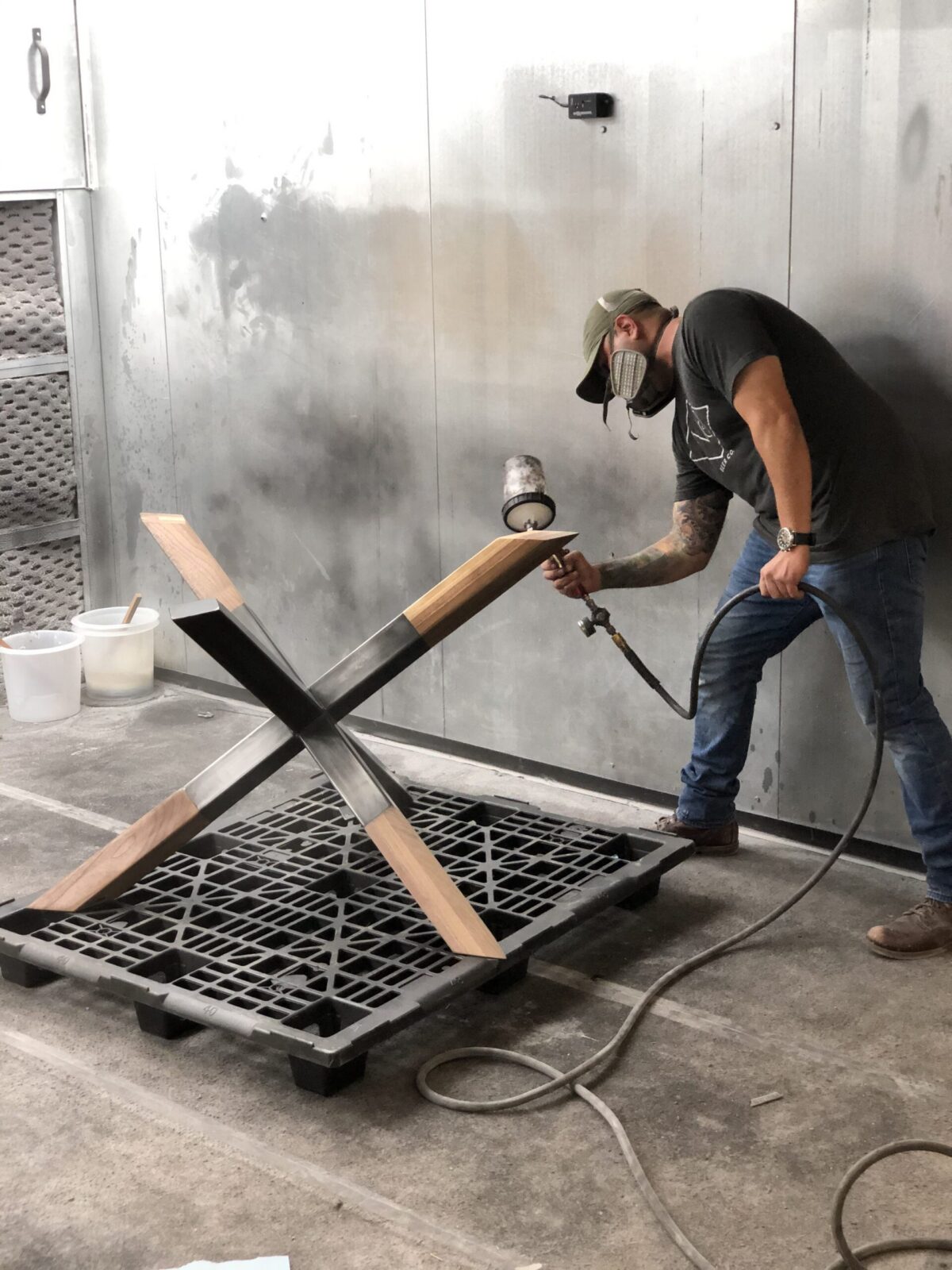 Craftsman applying finish to a wooden X-shaped table base using a spray gun in a workshop, wearing a mask and standing on a concrete floor.
