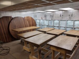 Multiple wooden rectangular tabletops, both rectangular and round, placed on wooden supports in a workshop with filtered ventilation.