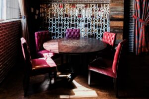 A wooden round tabletop with an intricate design at the center, surrounded by plush red leather chairs with button-tufted backs. The space has a warm ambiance with sunlight streaming through a nearby window. A decorative geometric divider wall is in the background, along with exposed brick walls and bold-patterned curtains.