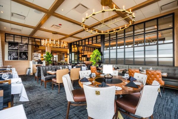 A luxurious restaurant interior featuring elegant modern furniture, large chandeliers, and expansive windows. The seating arrangement includes a mix of upholstered chairs, leather seats, and round wooden tables set with wine glasses and tableware.