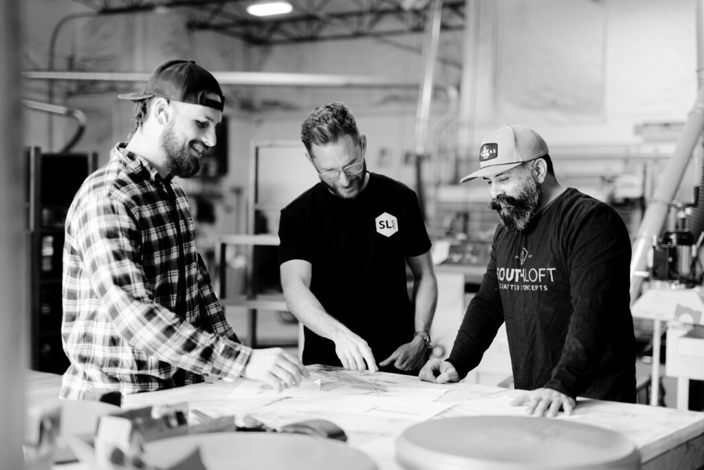 Three craftsmen from South Loft collaborate at a workbench, reviewing design plans in a workshop setting. The team is smiling, focused on the project, with one of them pointing at the plans. The atmosphere is casual yet professional, highlighting teamwork and attention to detail in the furniture-making process.