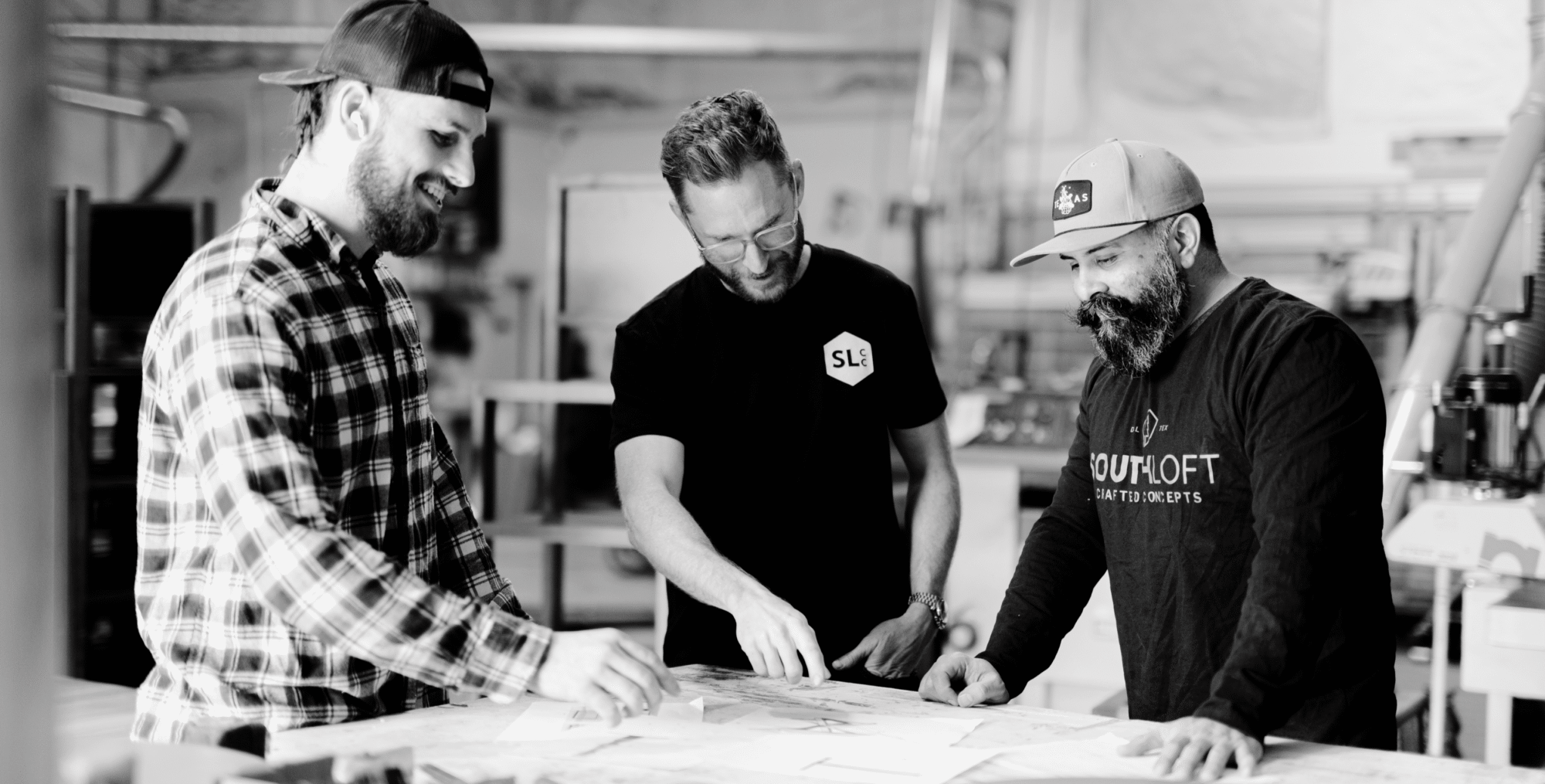 Three craftsmen from South Loft collaborate at a workbench, reviewing design plans in a workshop setting. The team is smiling, focused on the project, with one of them pointing at the plans. The atmosphere is casual yet professional, highlighting teamwork and attention to detail in the furniture-making process.