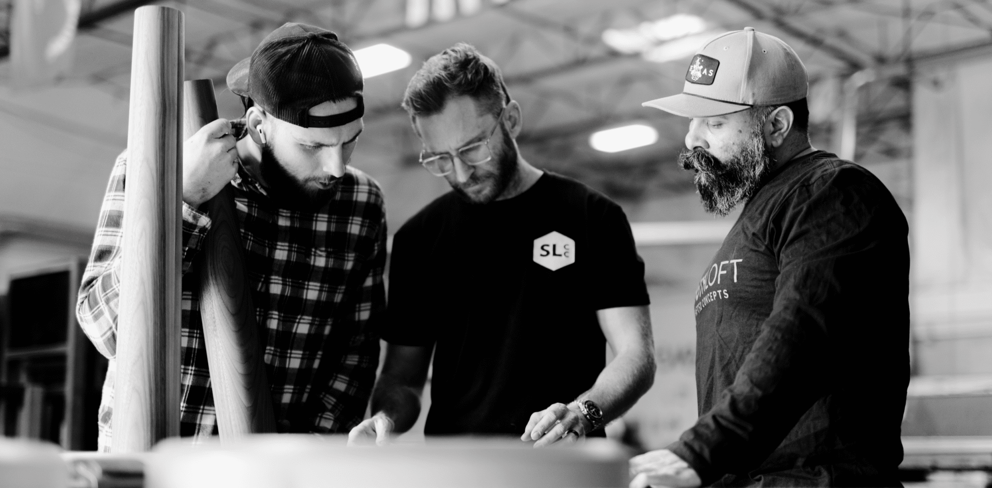 Three South Loft craftsmen examine design plans in a workshop setting. The image is in black and white, showing the team members deeply focused on a project. One holds a wooden component, while the others review the plans spread out on the table. The atmosphere reflects collaboration and precision in their craftsmanship.