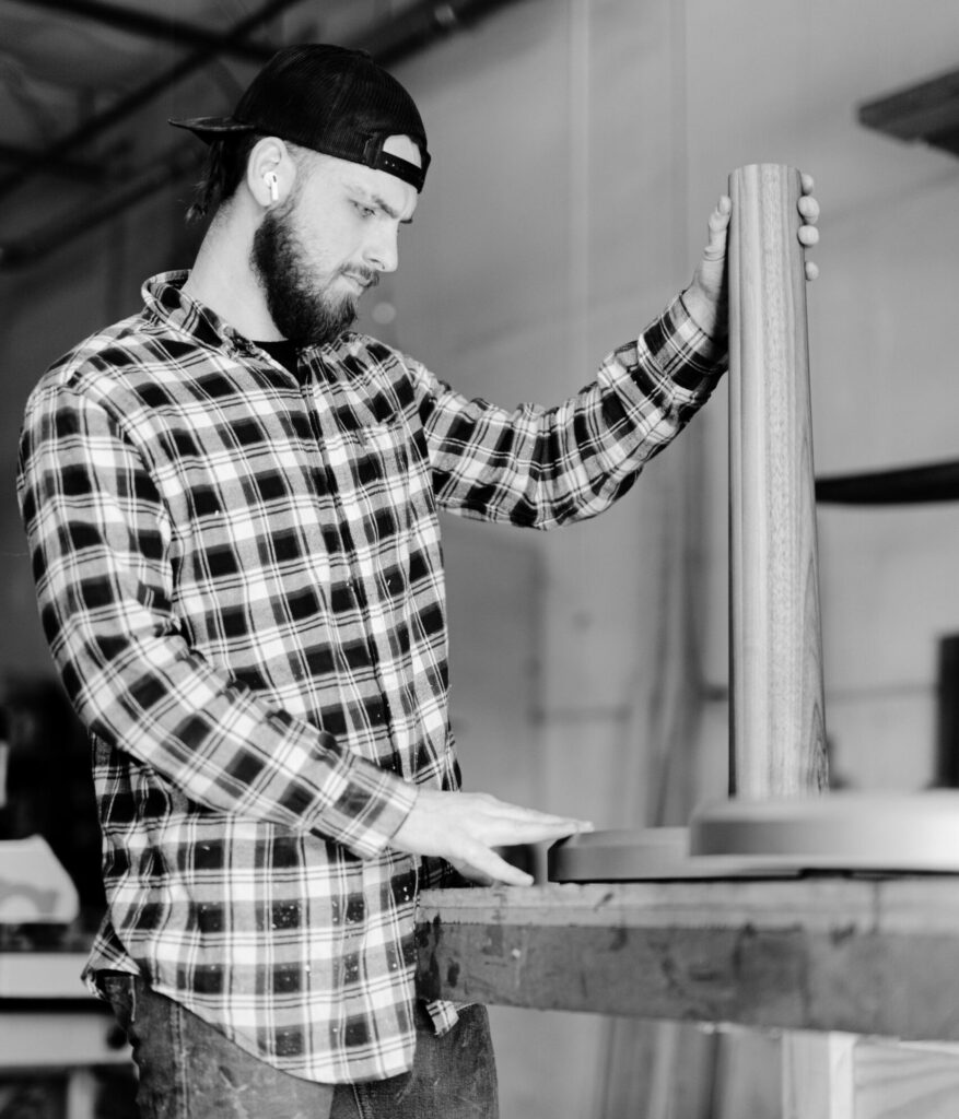 A craftsman in a plaid shirt and backward cap works with a wooden component in a workshop. He focuses intently on positioning a cylindrical piece on a round base, indicative of custom furniture creation.