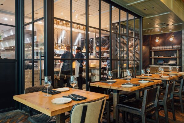 South Loft custom wooden tables and chairs in a modern restaurant with an open kitchen behind glass walls.
