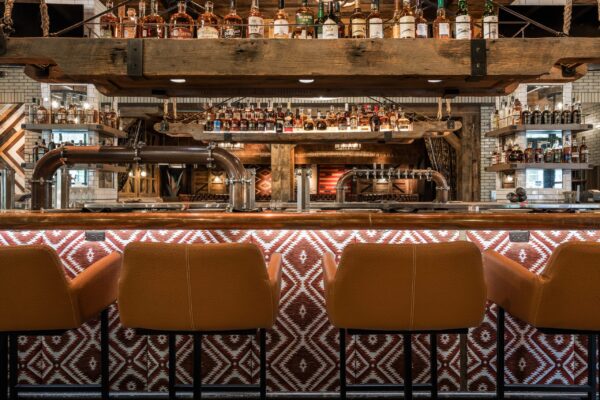 South Loft custom bar with leather stools and a wooden overhead shelf holding various liquor bottles.