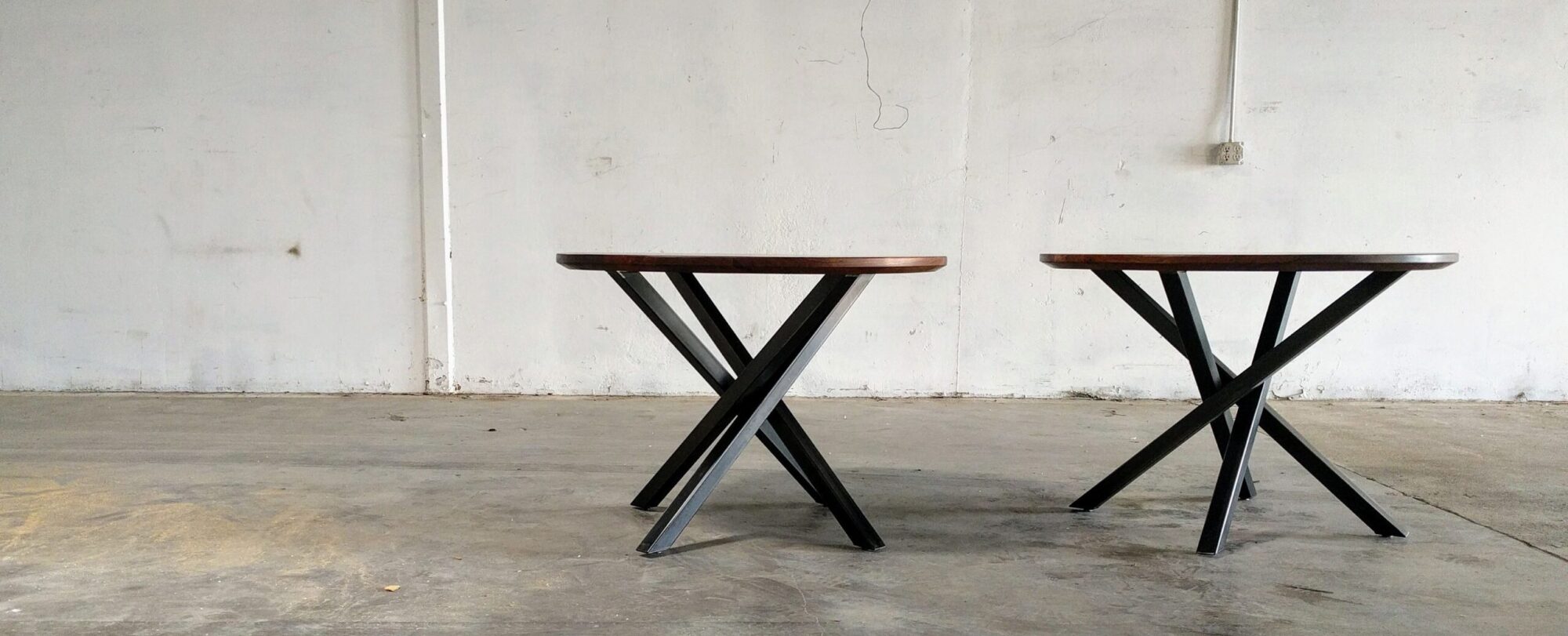Two round wooden tables with black metal crisscross legs designed by South Loft, placed against a plain white wall in a minimalist setting.