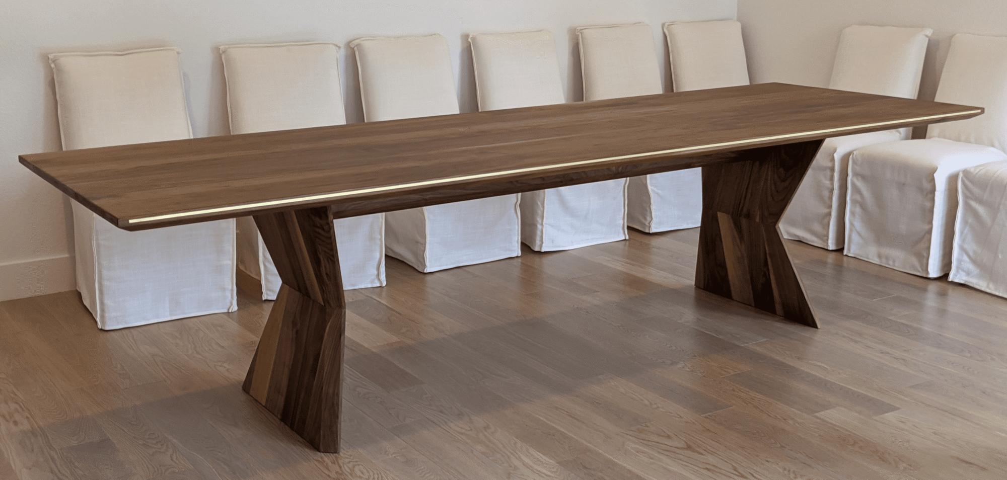 Custom walnut dining table with angular base designed by South Loft, placed in a dining room with white upholstered chairs.