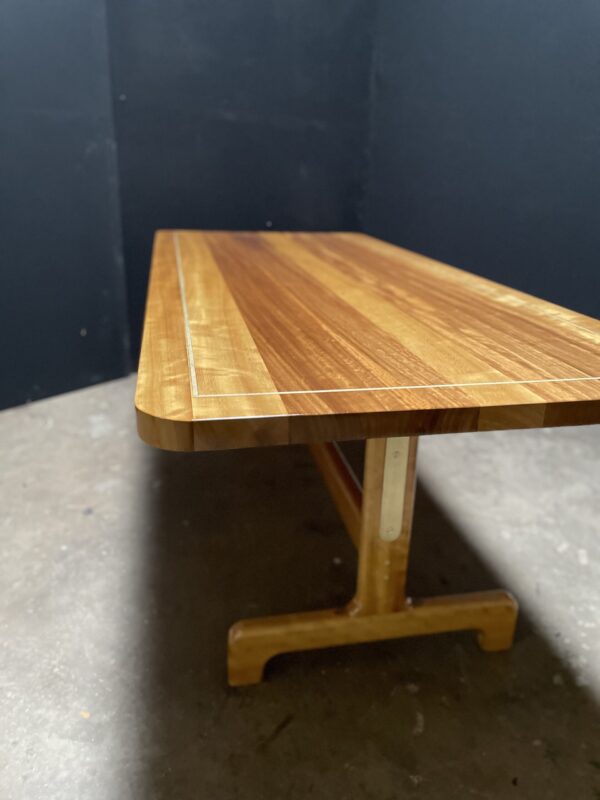 A sleek custom wooden dining table by South Loft with rounded edges and a trestle base, highlighted by a natural wood grain finish and brass accents.