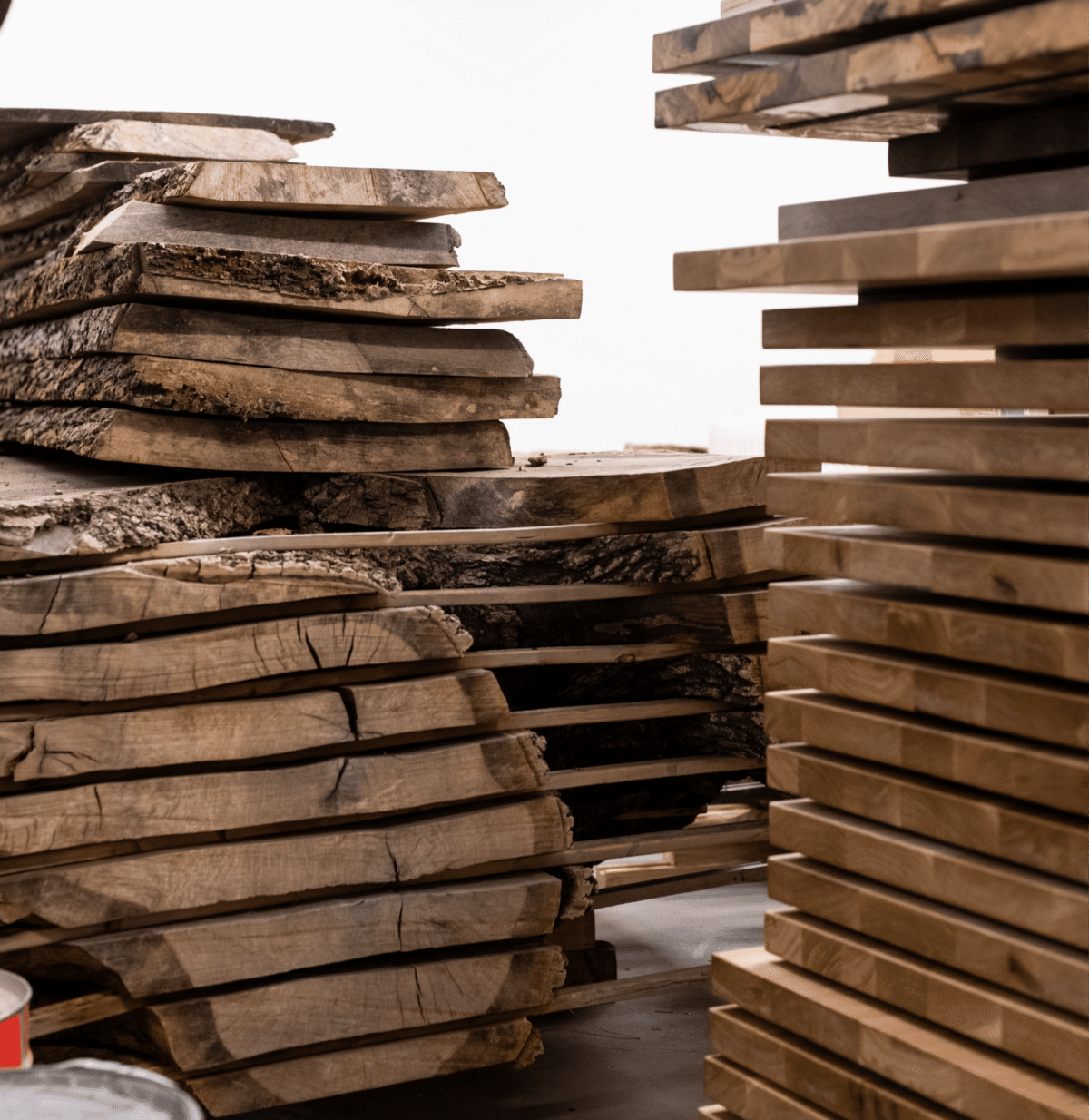 Stacks of rough and processed wooden slabs are arranged in a workshop, showcasing different stages of wood preparation for custom furniture.