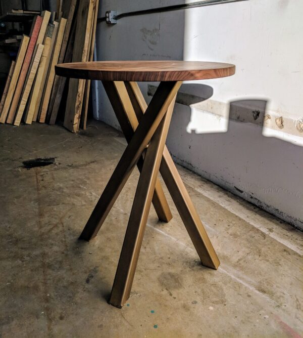 A round wooden side table by South Loft with a natural wood finish and intersecting metal legs, positioned in a workshop space with wood planks stacked in the background.