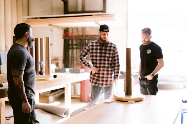 Three men, two standing around a workshop table with wooden materials, are discussing a project in a well-lit woodworking shop. The workshop is branded with South Loft's logo in the background.