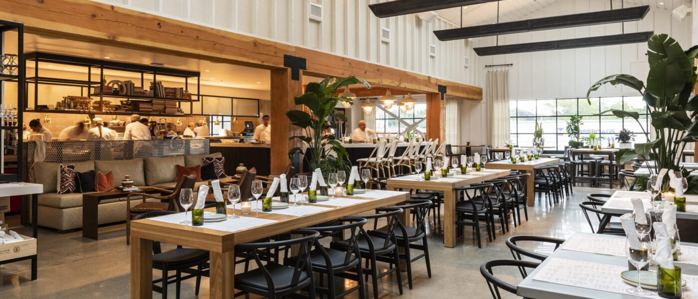 A bright, modern restaurant with open kitchen space, communal wooden tables, and black chairs. The tables are set with wine glasses and napkins, and lush green plants add to the inviting ambiance. A team of chefs works in the open kitchen in the background.