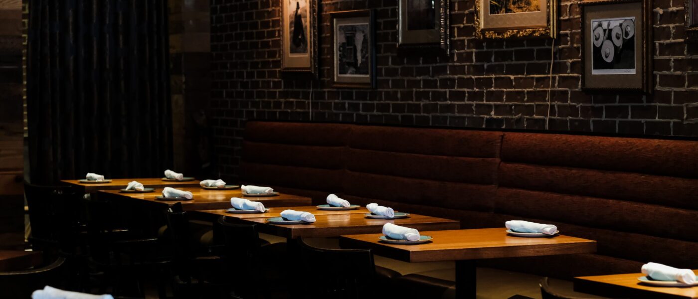 A dimly lit dining area with exposed brick walls, featuring wooden tables set with rolled napkins and plates, complemented by framed photographs and artwork on the walls.