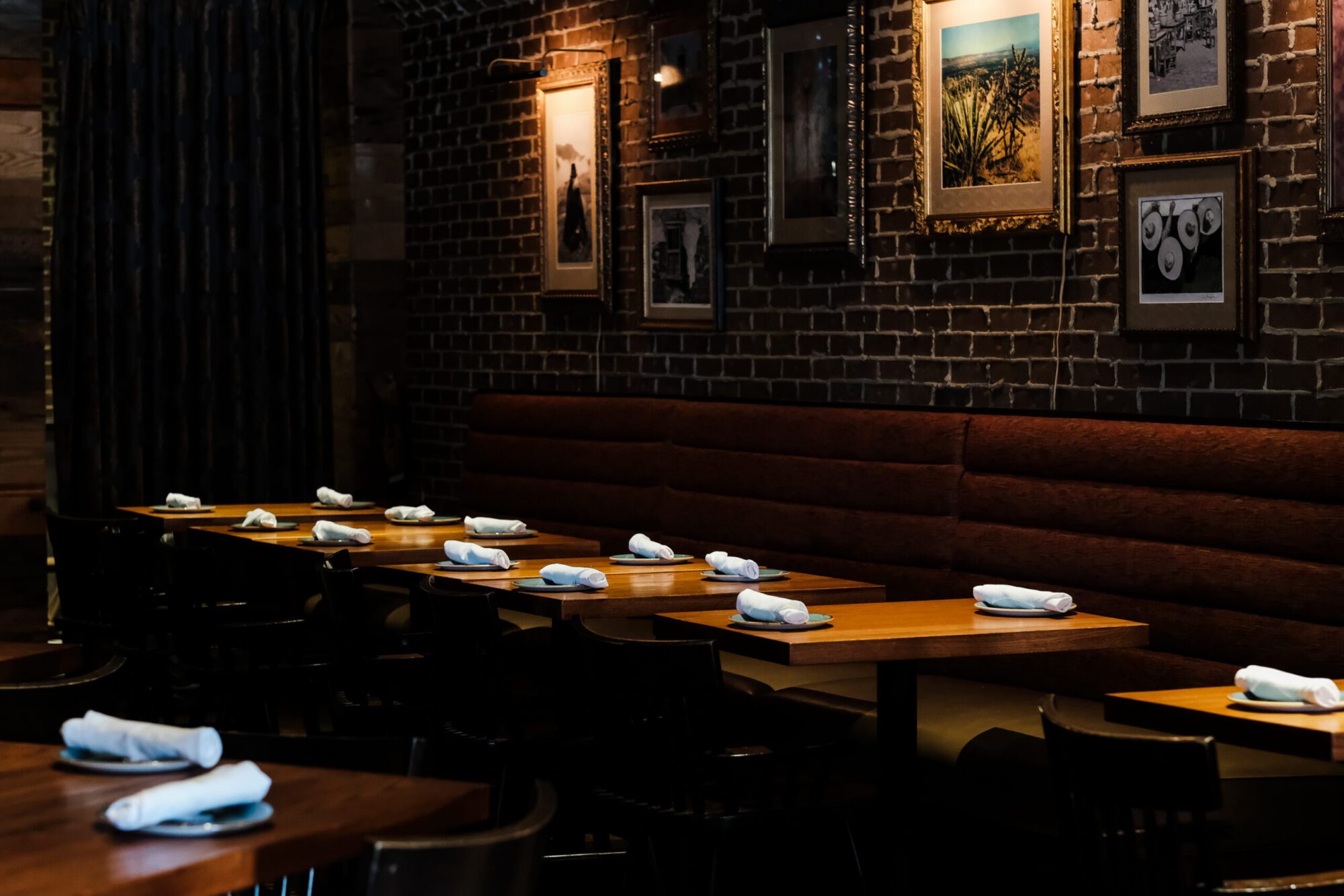 A dimly lit dining area with exposed brick walls, featuring wooden tables set with rolled napkins and plates, complemented by framed photographs and artwork on the walls.
