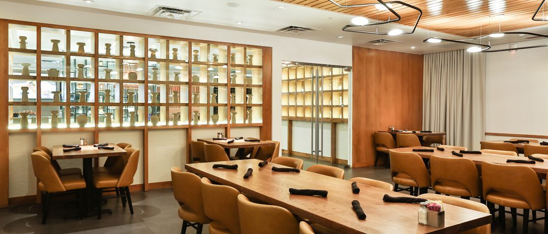 A modern restaurant dining area featuring wooden tables, tan leather chairs, and a backlit shelf display with decorative items. The ceiling has unique light fixtures and wooden paneling, adding warmth to the space.