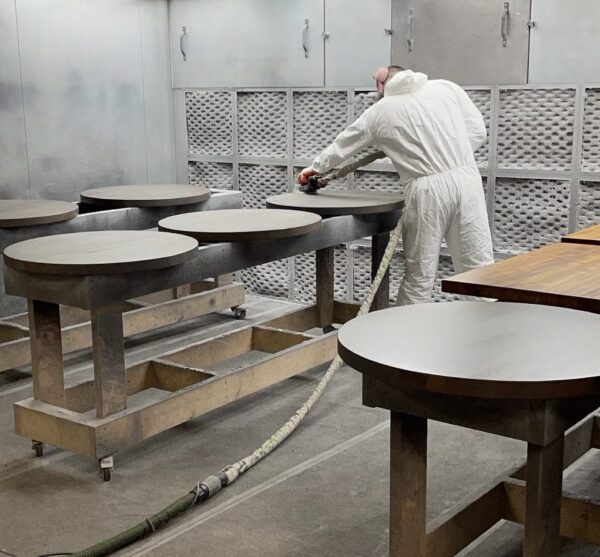 A worker in protective gear sanding round tabletops in a finishing booth at South Loft's production facility.