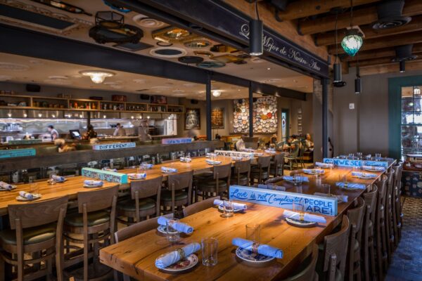 A long wooden dining table, custom-made by South Loft, set with plates, glasses, and rolled napkins, situated in a vibrant restaurant with an open kitchen, showcasing both comfort and craftsmanship.