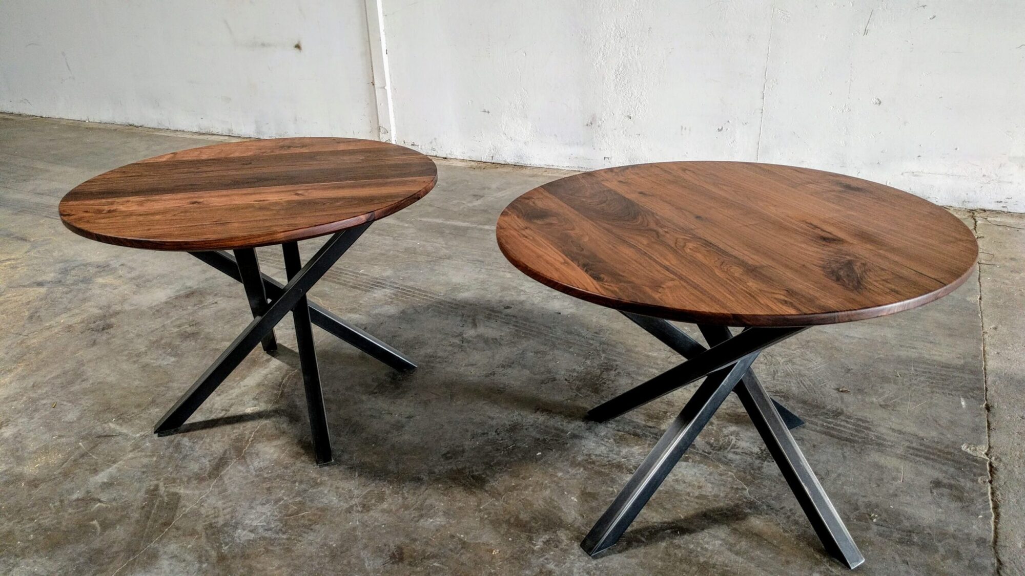 Two 120 Series round tabletop walnut tables with crossed metal legs placed on a smooth concrete floor. The tables have a warm, natural wood grain finish and modern, black metal bases, showcasing SouthLoft’s craftsmanship.