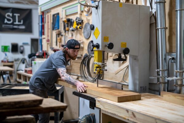 A South Loft craftsman operates a large woodworking machine, cutting a piece of wood in a workshop, demonstrating the precision and care South Loft applies to its handcrafted furniture.