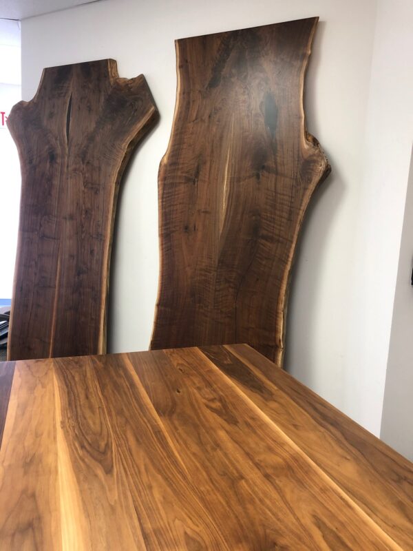 Two large live edge walnut slabs stand against a white wall, with a matching finished walnut tabletop in the foreground, showcasing natural wood grain patterns.