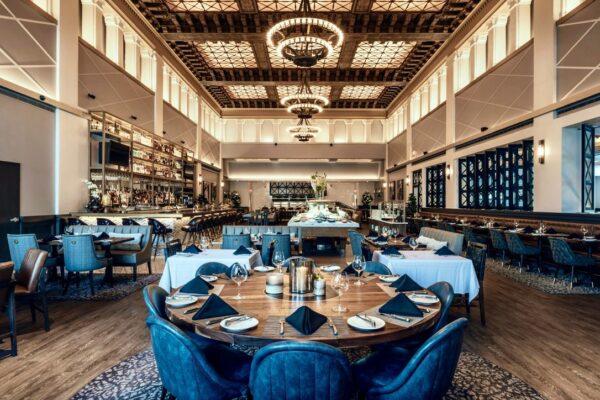 A grand dining hall with high vaulted ceilings, elegant chandeliers, and a lavish circular dining table at the forefront, crafted by South Loft, surrounded by plush blue chairs in an upscale restaurant setting.
